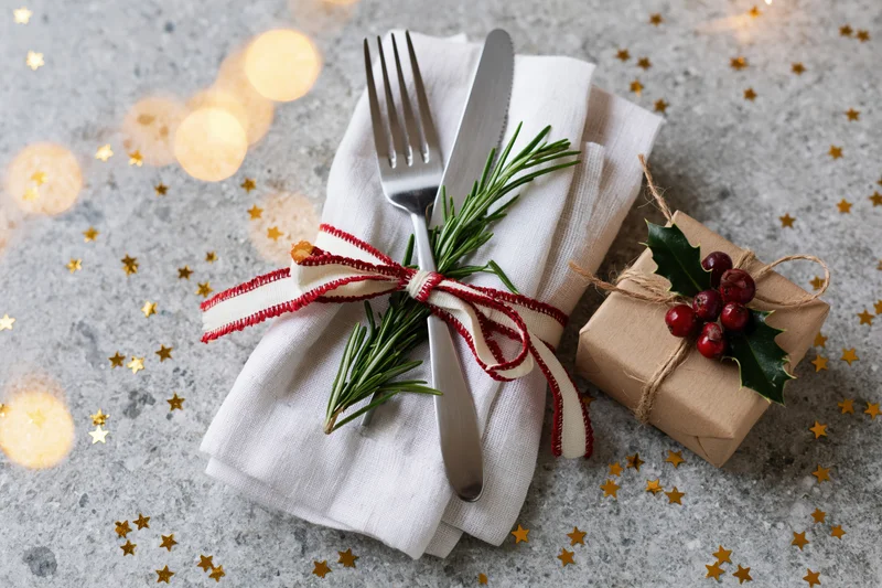 Fork and knife wrapped in a white linen napkin tied with a red and white ribbon, placed beside a small gift wrapped in brown paper with holly and berries, on a gray surface scattered with gold stars and soft glowing lights