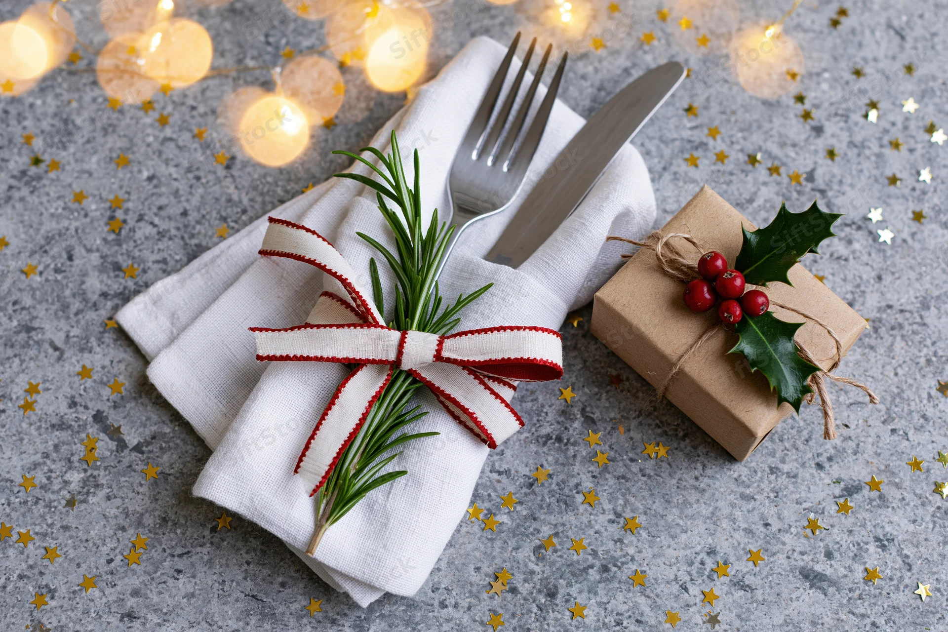Fork and knife wrapped in a white linen napkin with rosemary sprig and red ribbon bow beside a small gift box decorated with holly and berries on a gray surface with golden stars and warm string lights