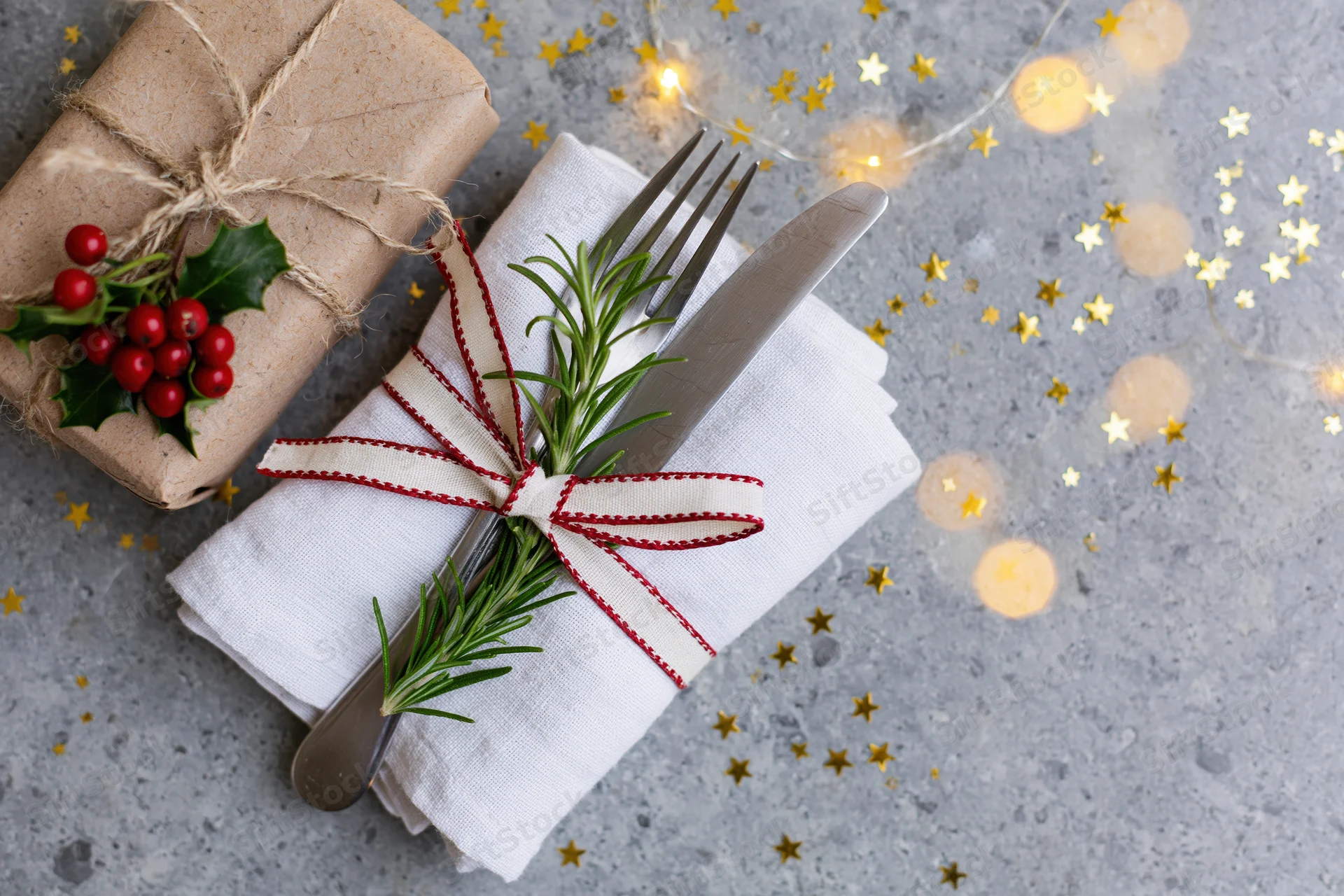 Fork and knife wrapped in a white napkin with rosemary and red ribbon, beside a holly-decorated gift on a gray surface with golden stars and fairy lights