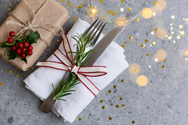 Fork and knife wrapped in a white napkin with rosemary and red ribbon, beside a holly-decorated gift on a gray surface with golden stars and fairy lights