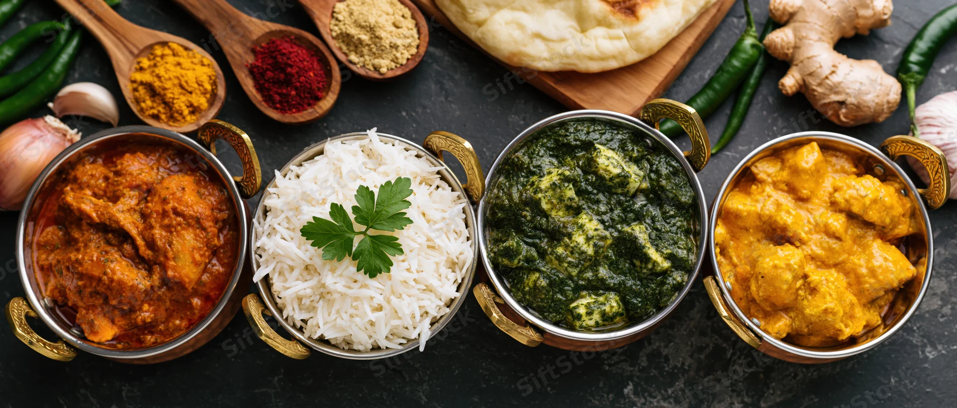 Four indian curry dishes in brass-handled karahi bowls with rice and spices arranged on dark surface