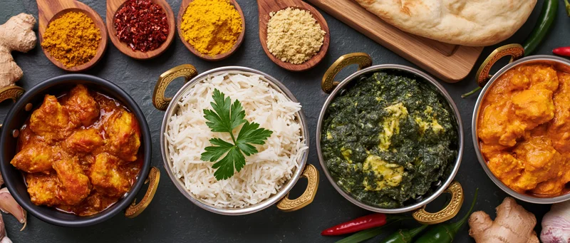 Four indian curry dishes in brass-handled karahi bowls with rice and naan on dark surface