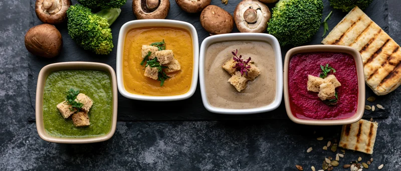 Four ceramic bowls of cream soup arranged in a row on a dark surface with broccoli, mushrooms, croutons and garnish
