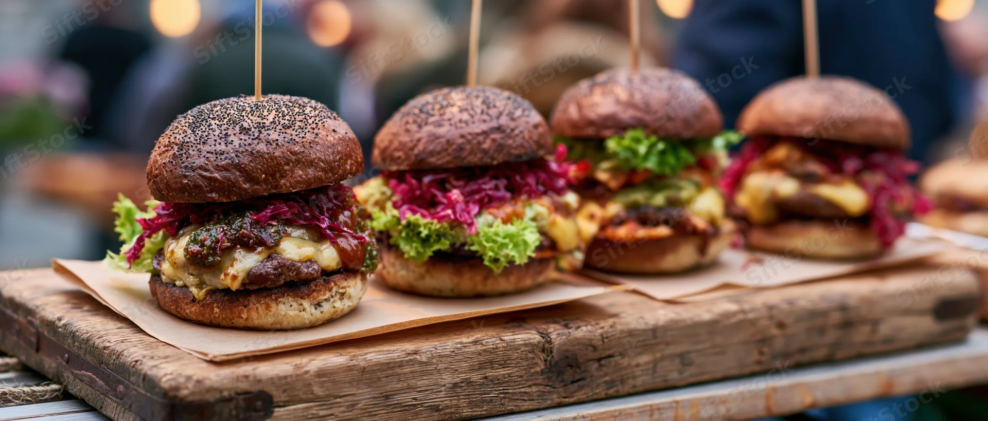 Four gourmet burgers with dark poppyseed buns lined up on a wooden tray outdoors at evening