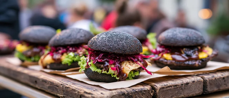 Four gourmet burgers with dark poppy-seed buns lined up on a wooden tray