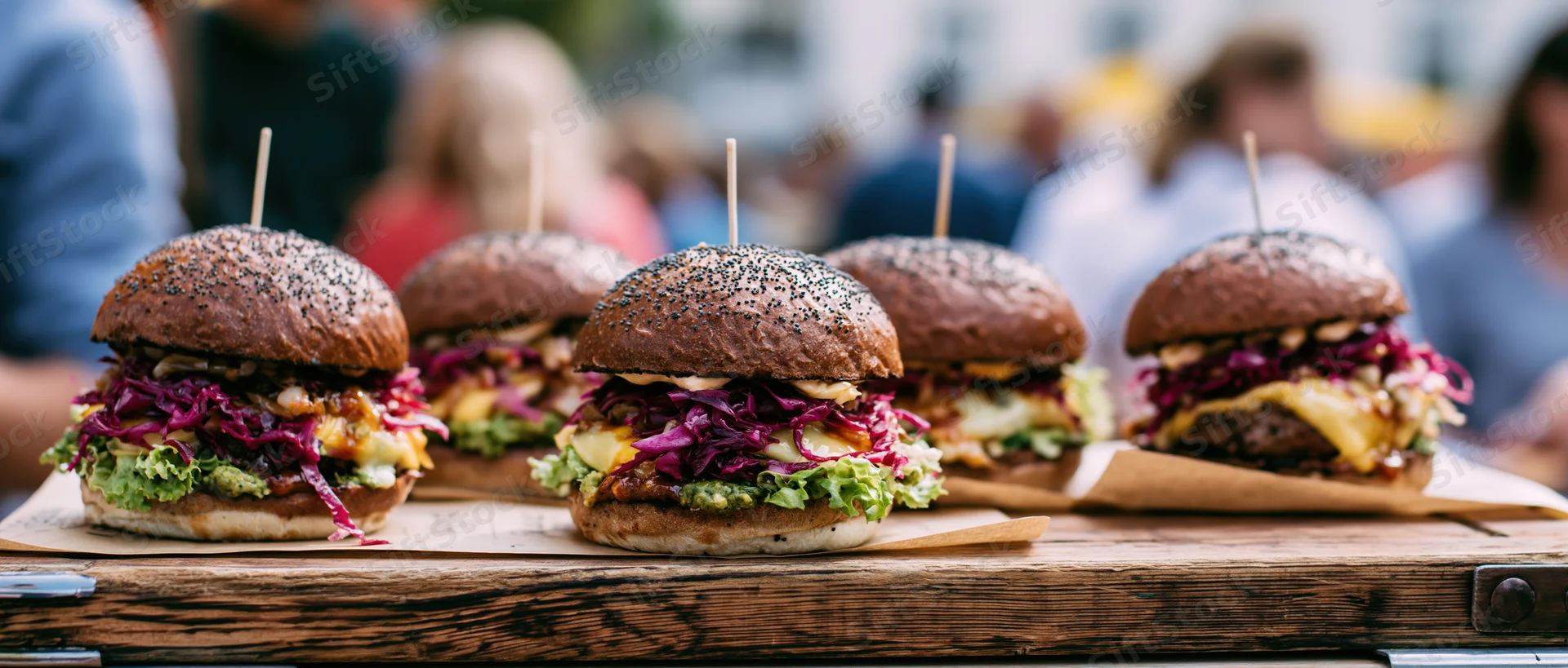 Four gourmet burgers with dark poppy-seed buns lined up on a wooden tray outdoors at a food market