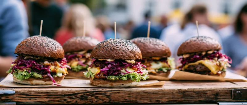 Four gourmet burgers with dark poppy-seed buns lined up on a wooden tray outdoors at a food market