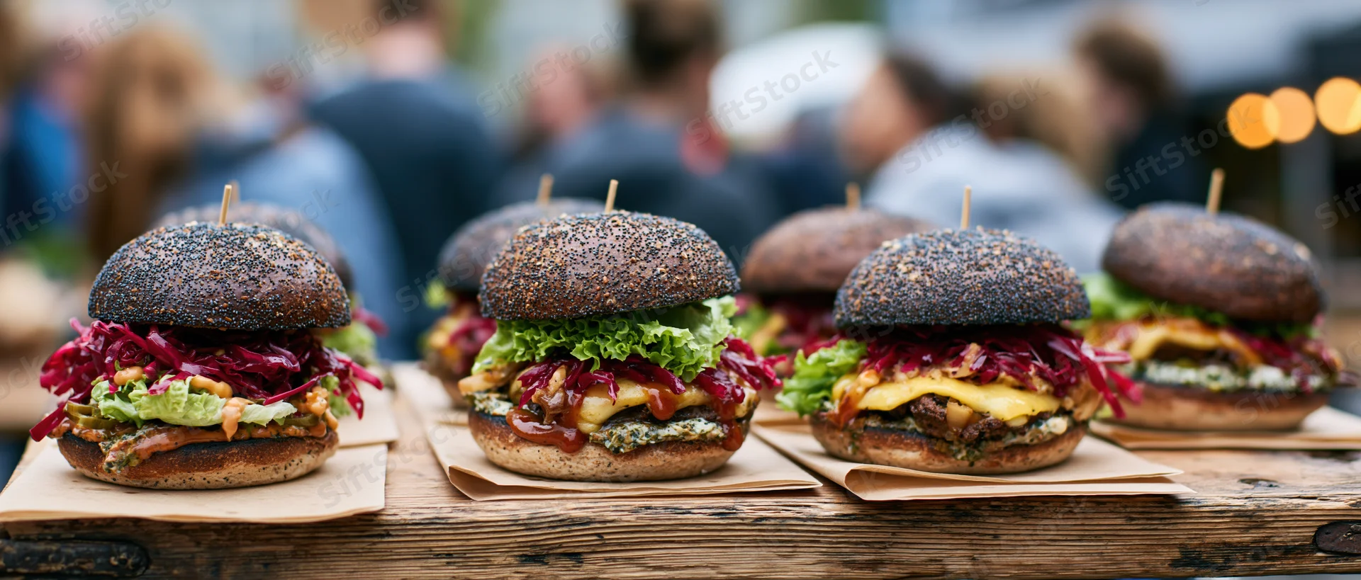 Four gourmet burgers with dark poppy-seed buns lined up on a wooden table outdoors