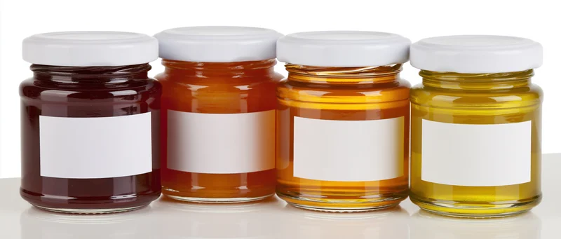 Four glass jars of honey with white lids and blank labels in a row on a white surface