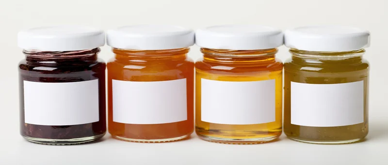 Four glass jars with white lids and blank labels lined up in a row on a white surface