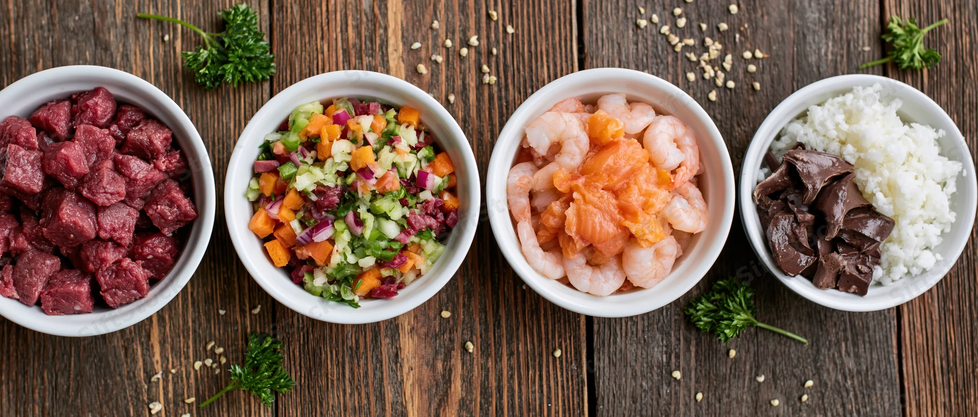 Four white bowls with beef, shrimp, salmon, rice, chocolate, and salad arranged in a row on a wooden table