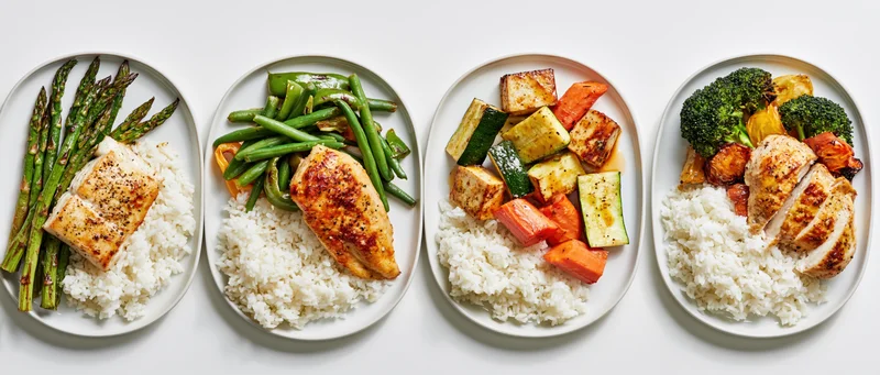 Four plates with rice, vegetables, and protein arranged side by side in a flat lay