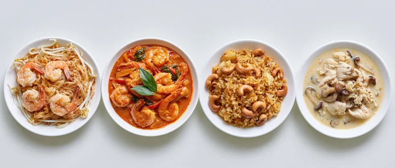 Four thai dishes served in white bowls arranged in a row on a white surface