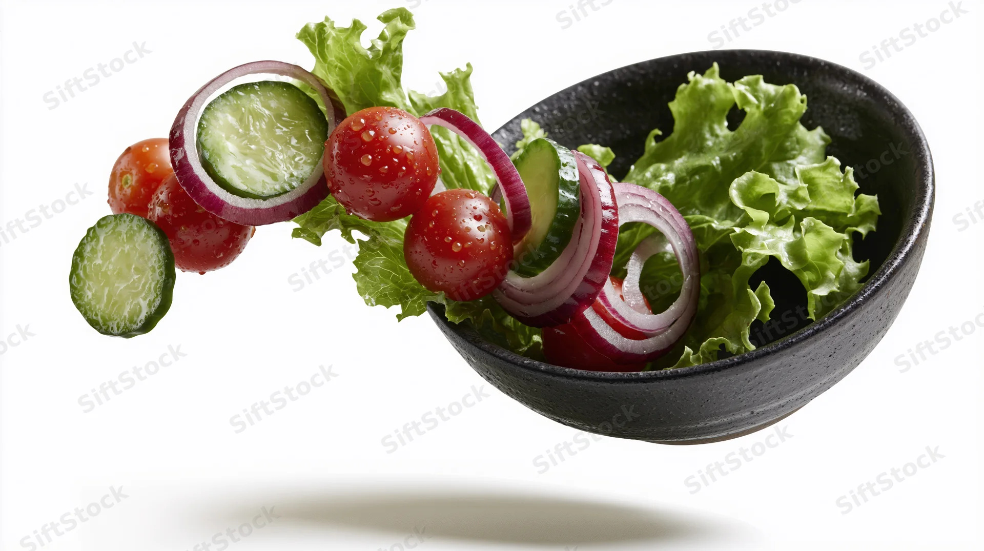 Fresh salad ingredients falling into a dark ceramic bowl against white background