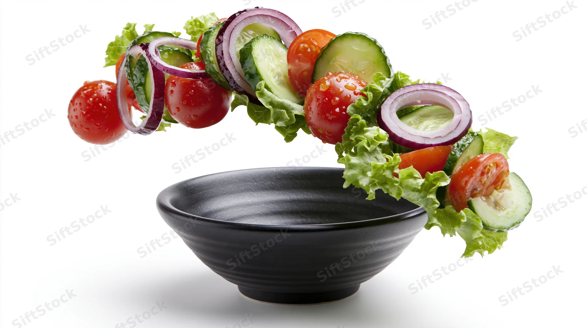 Fresh salad ingredients falling into a dark ceramic bowl against white background