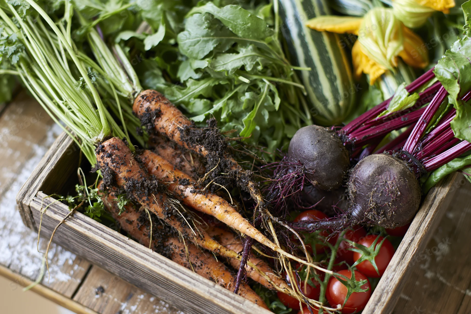 Fresh harvested vegetables including carrots, beets, tomatoes, and zucchini arranged in a shallow wooden crate