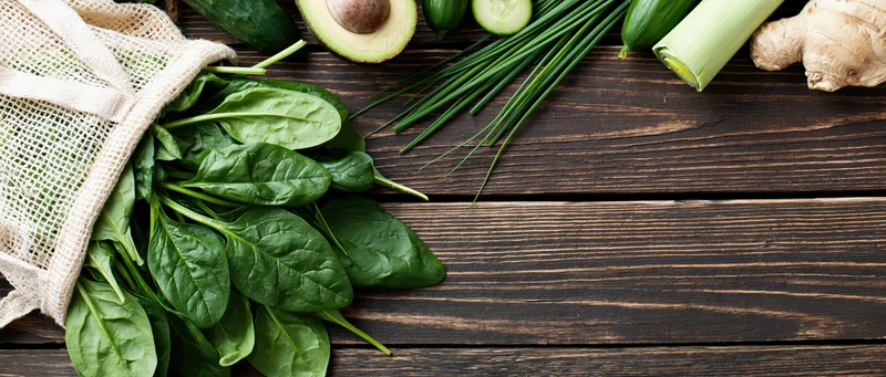 Fresh spinach spilling from a reusable mesh bag onto a dark wooden table with vegetables arranged around it