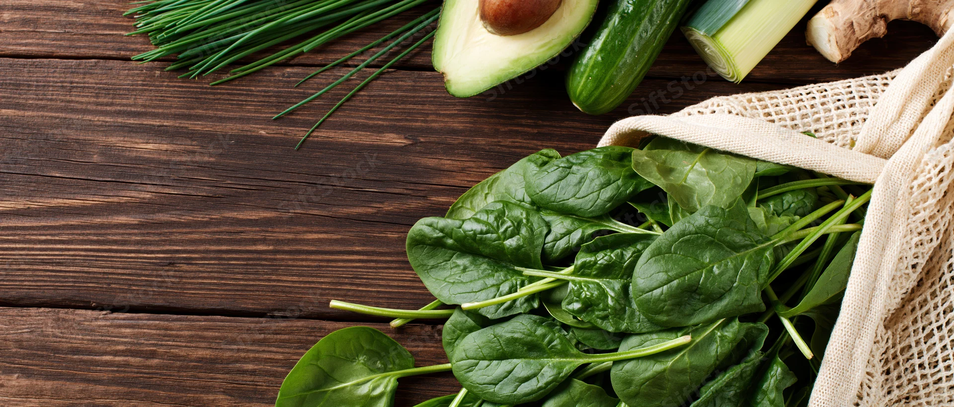 Fresh spinach leaves spilling from a cotton mesh bag onto a wooden table with other vegetables nearby