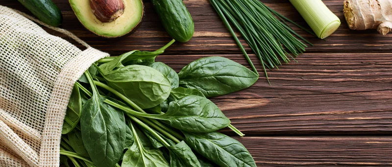 Fresh spinach leaves spilling from a cotton mesh bag onto a wooden table with other vegetables