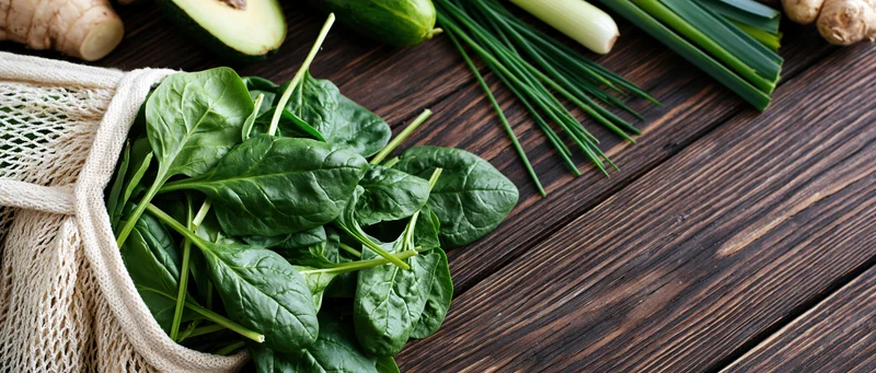 Fresh spinach leaves spilling from a cotton mesh bag on a wooden table with other vegetables