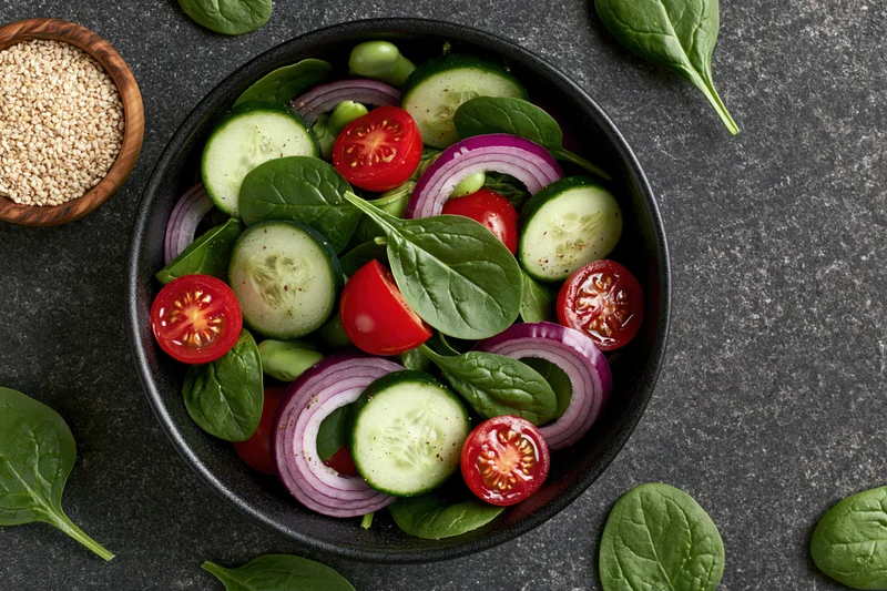 Fresh vegetable salad in a black ceramic bowl with scattered spinach leaves and sesame seeds on a dark stone surface, top view