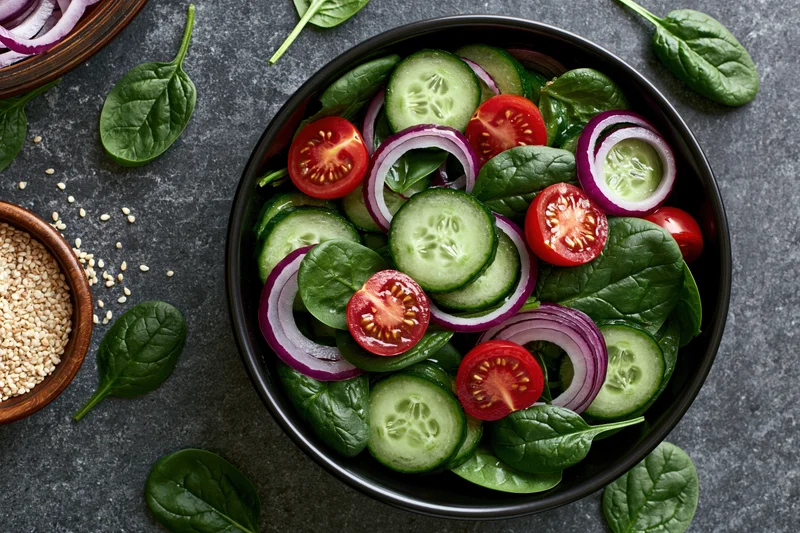Fresh vegetable salad in a black ceramic bowl viewed from above on a dark countertop with scattered spinach leaves and sesame seeds nearby
