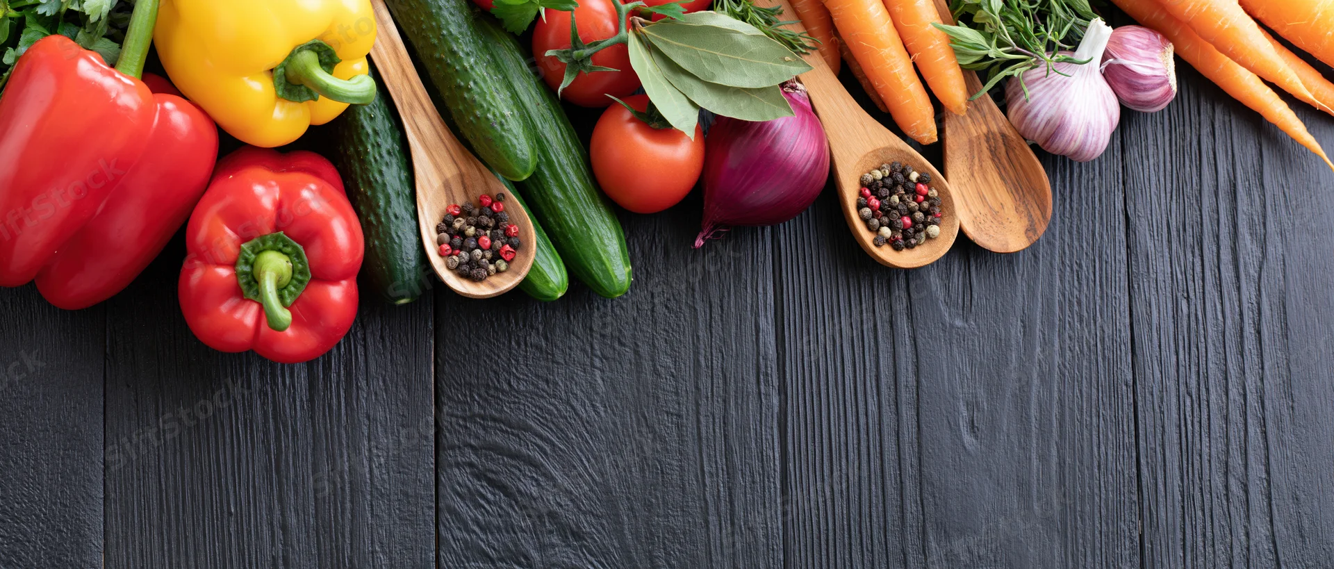 Fresh vegetables and spices arranged along the top edge of a dark wooden surface with copyspace below