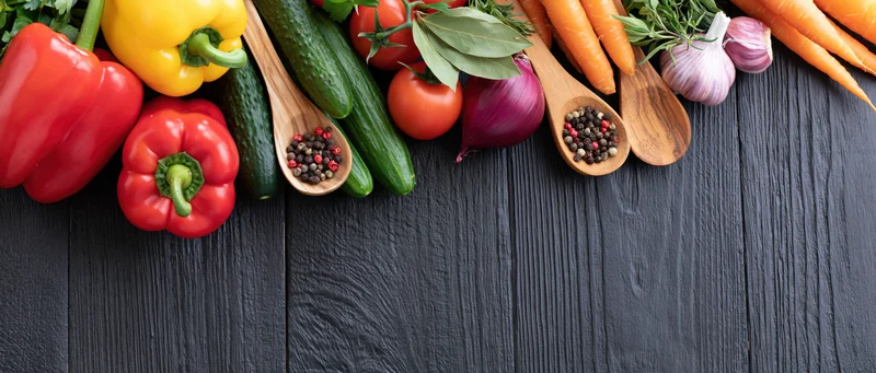 Fresh vegetables and spices arranged along the top edge of a dark wooden surface with copyspace below