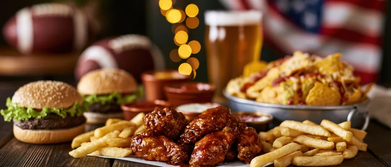 Game day spread of burgers, fries, wings, and beer on a dark wooden table with footballs and american flag in background