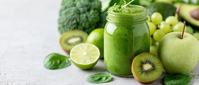 Thick green smoothie in a glass mason jar surrounded by fresh fruits and vegetables on a light countertop