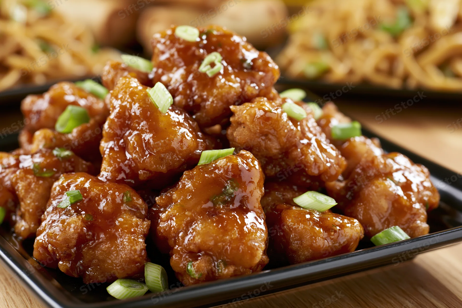 Glazed orange chicken pieces piled on a black square plate on a wooden table