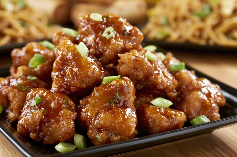 Glazed orange chicken pieces piled on a black square plate on a wooden table