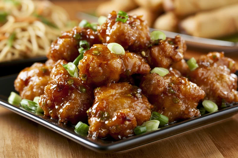 Glazed orange chicken pieces piled on a black square plate with green onion garnish on a wooden table