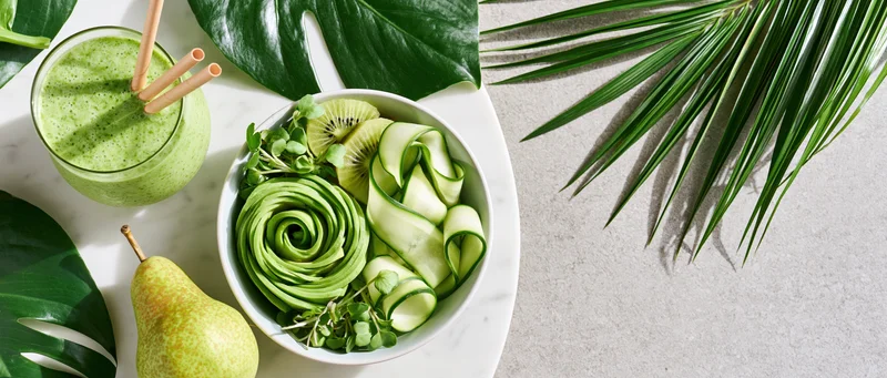 Green buddha bowl with avocado rose, kiwi, cucumber, and pear on marble surface with tropical leaves