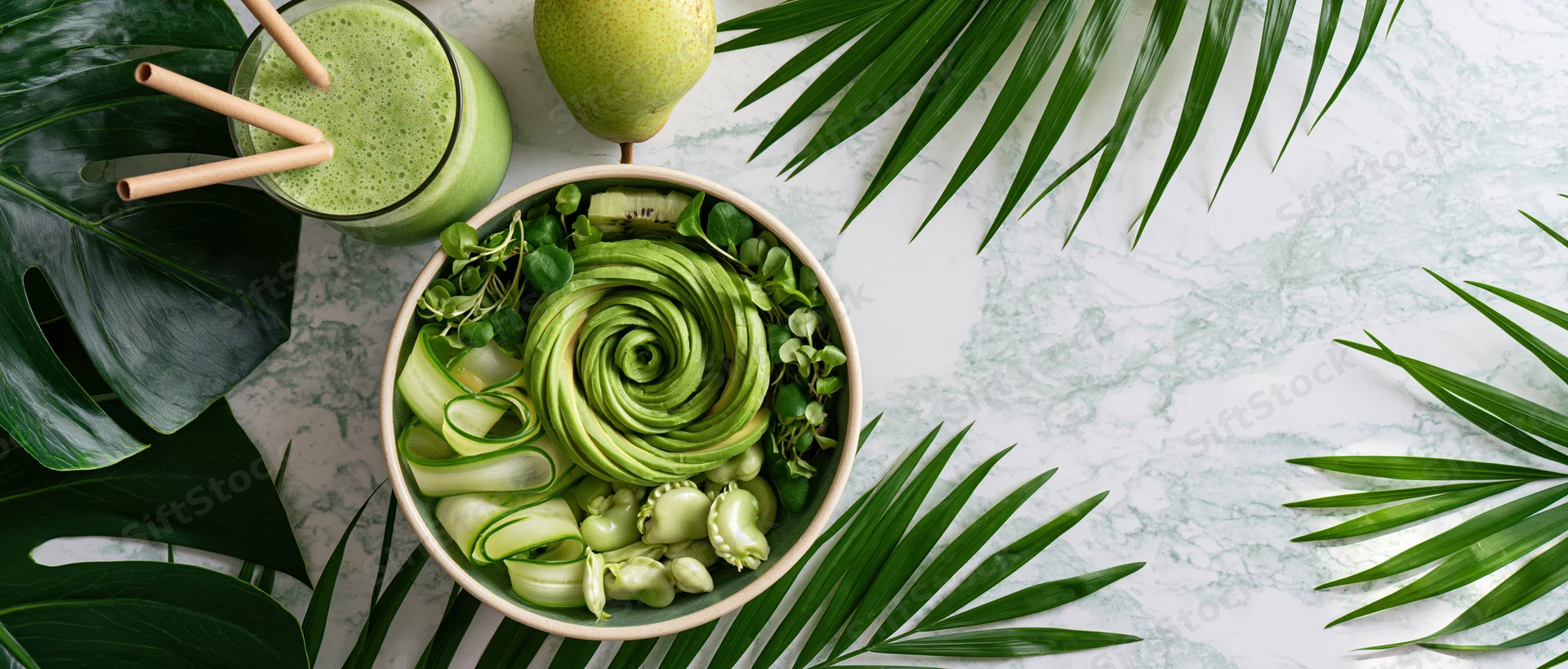 Green buddha bowl with avocado rose, kiwi, cucumber, and pear on marble surface surrounded by tropical leaves