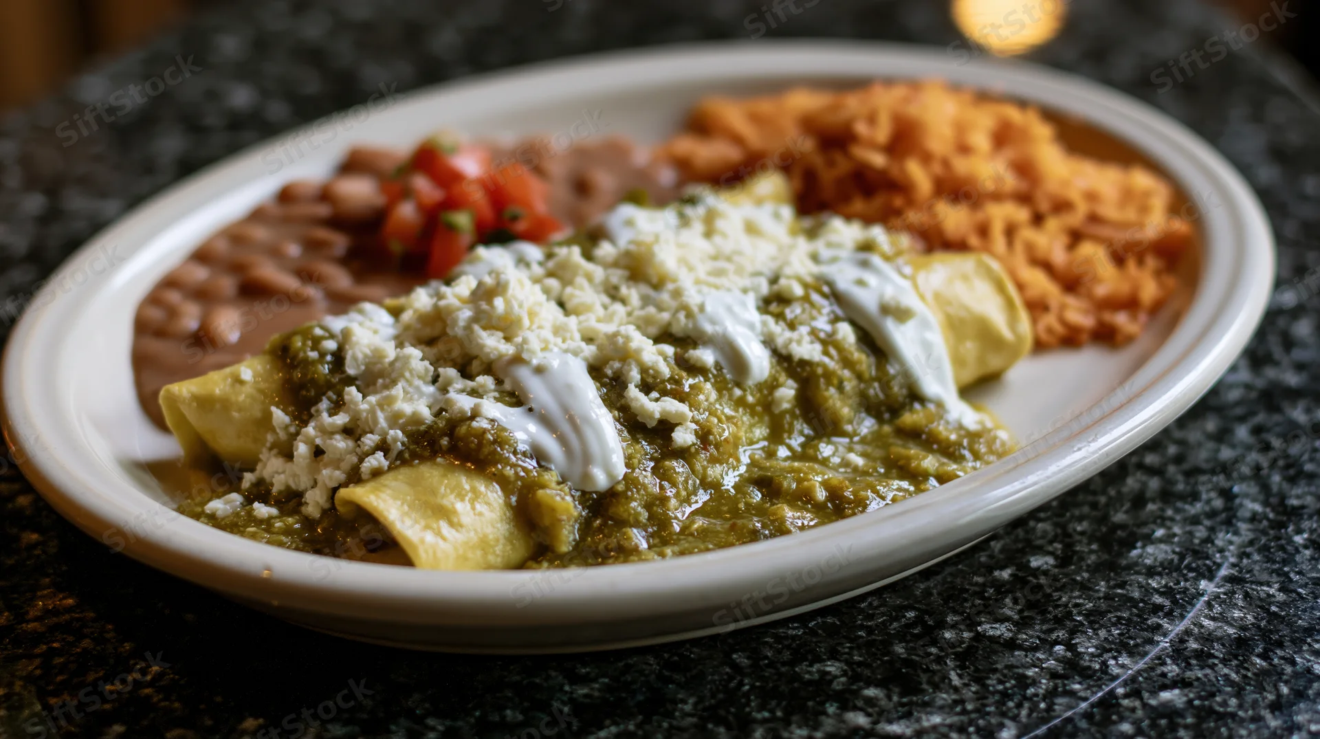 Green enchiladas smothered in tomatillo salsa verde with cheese and cream on a white oval plate with rice and beans on a granite table
