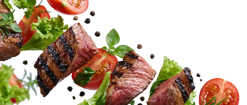 Grilled beef steak slices and fresh vegetables suspended midair against white background