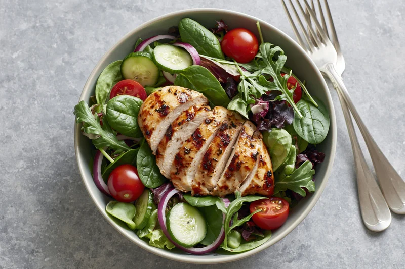Grilled chicken breast sliced and fanned over fresh garden salad in a bowl with vegetables and silverware on a gray surface