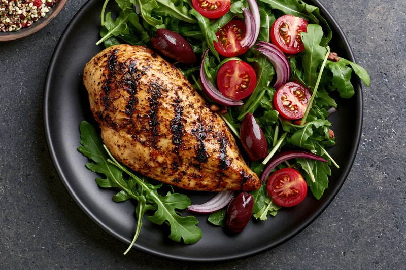 Grilled chicken breast with char marks served on a bed of arugula salad with tomatoes, red onions, and olives on a black plate