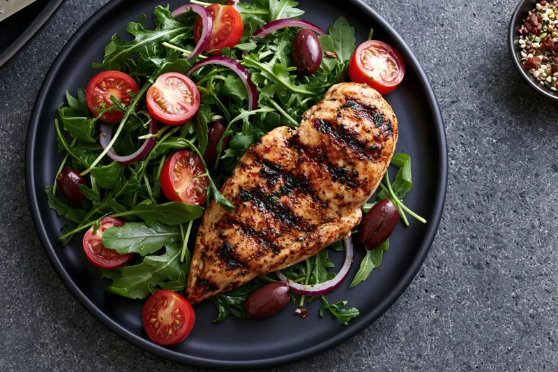 Grilled chicken breast with char marks on a bed of arugula salad with tomatoes, red onions, and olives on a black plate