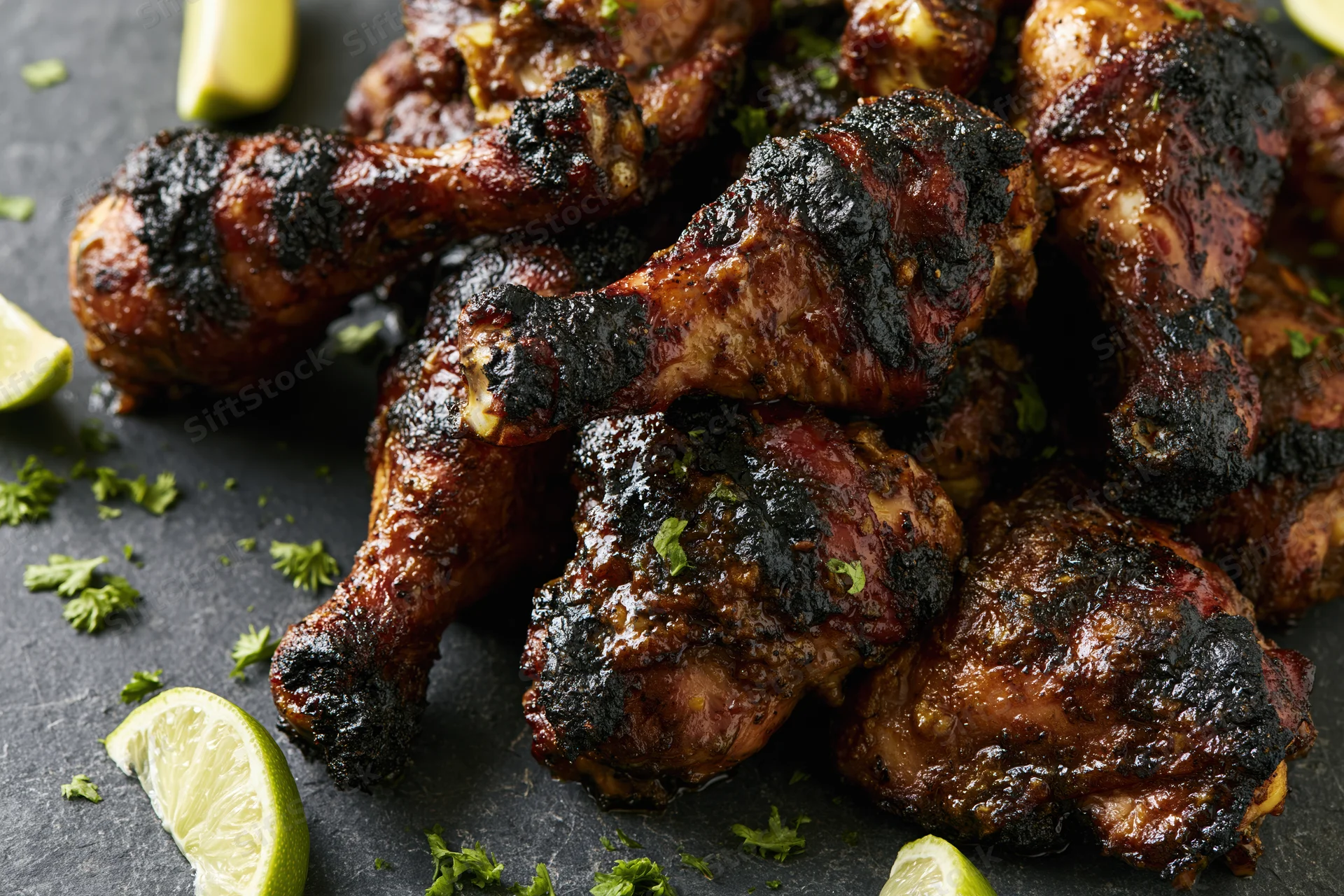 Grilled jerk chicken drumsticks and thighs piled on dark slate with lime wedges and cilantro garnish