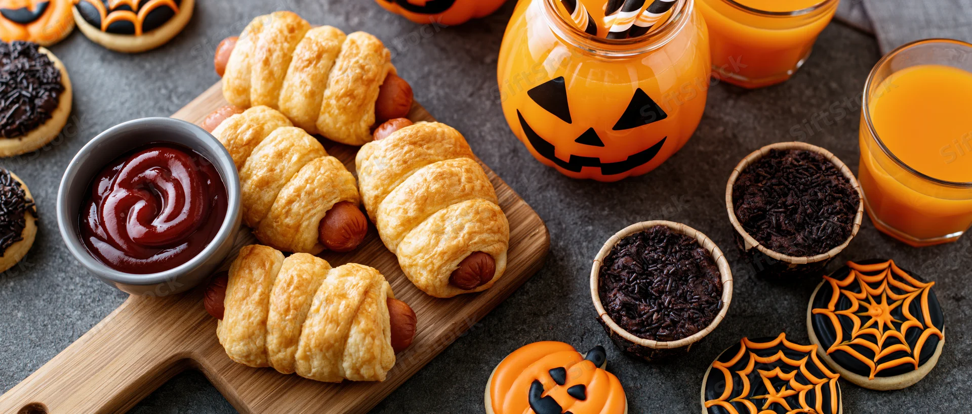 Halloween party food spread with pigs in a blanket, pumpkin juice, and themed cookies on dark stone surface