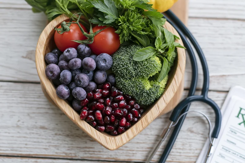 Heart-shaped wooden bowl filled with fresh vegetables and fruits on a wooden table with a stethoscope and prescription nearby