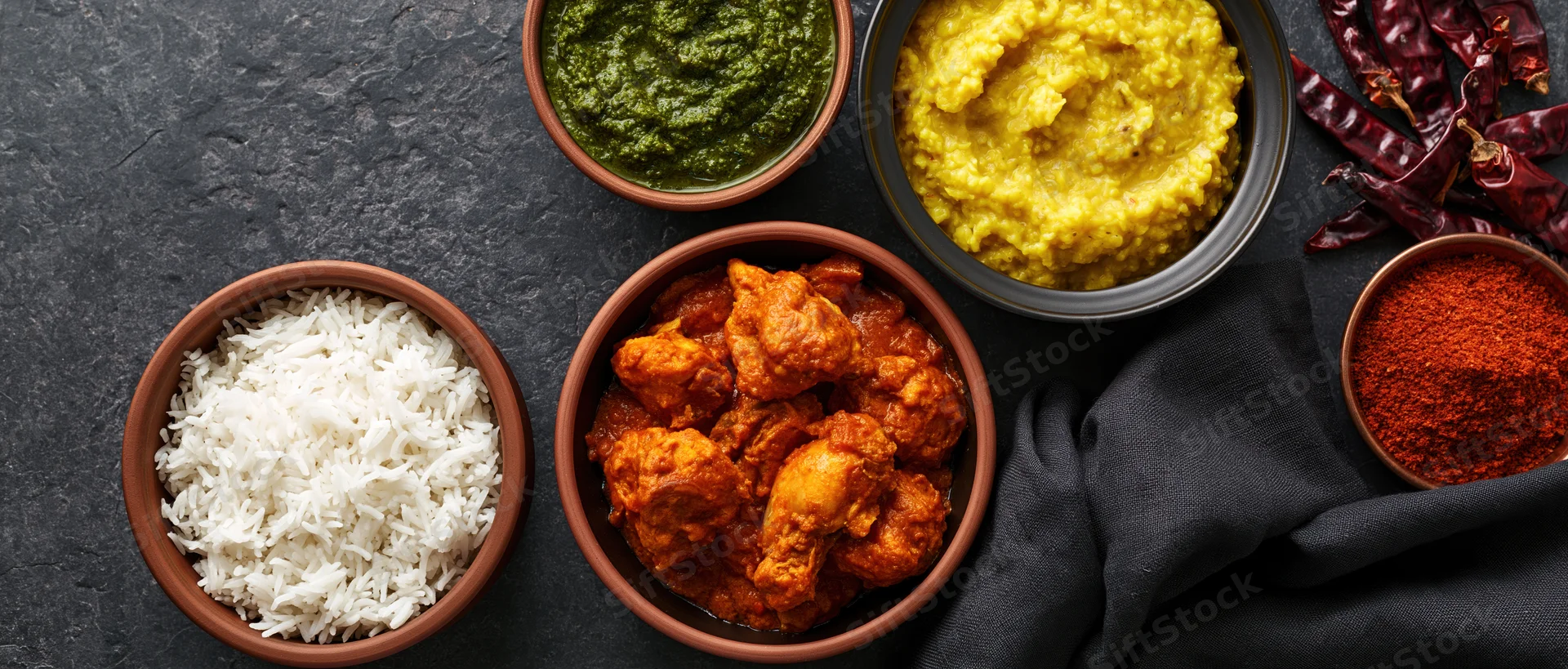 Indian meal spread with curry, rice, dal, chutney and spices arranged on dark slate surface overhead