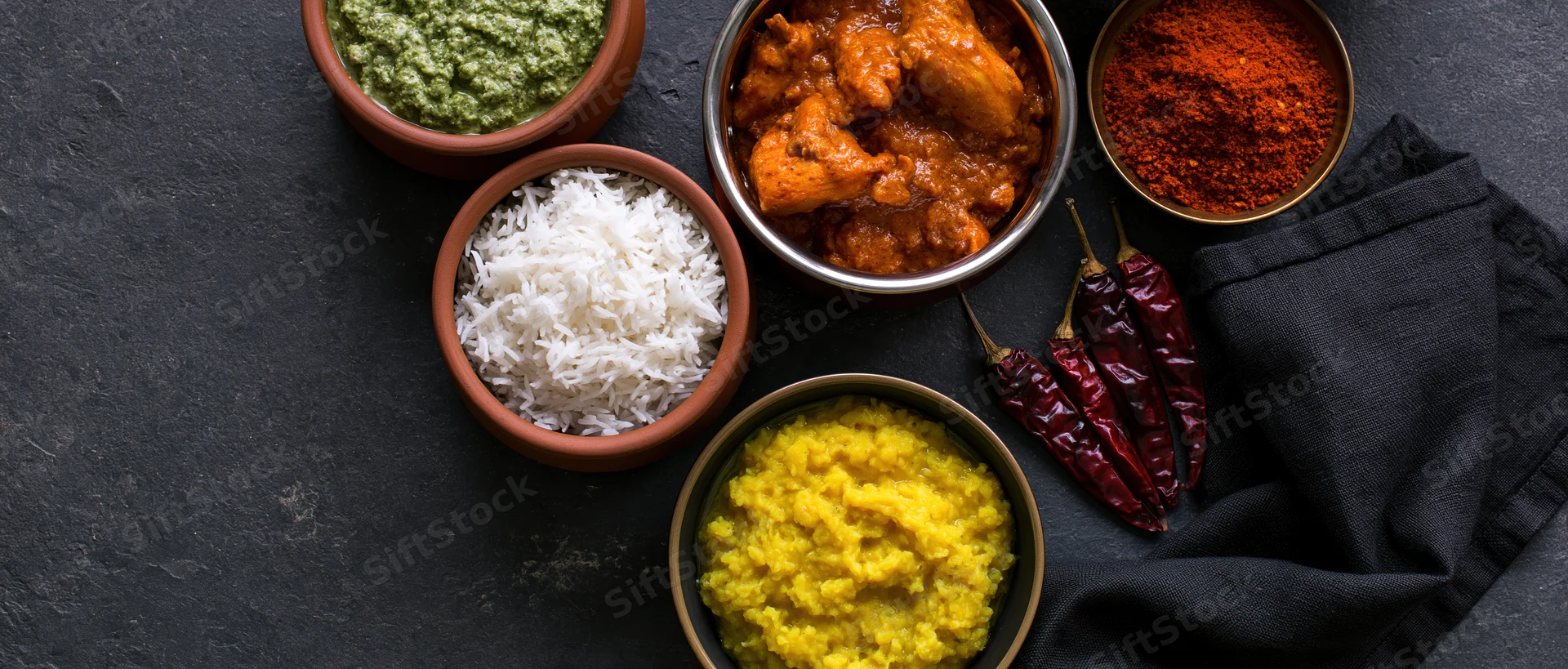 Indian meal spread with curry, rice, dal, chutney and spices arranged on dark slate surface overhead
