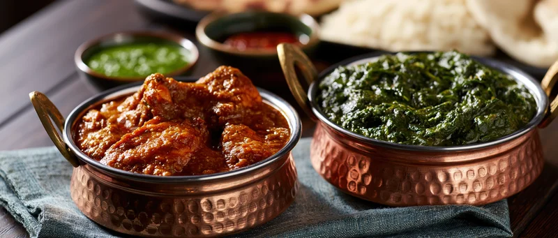 Copper bowls of curry and palak paneer served on a dark wooden table with naan and chutneys