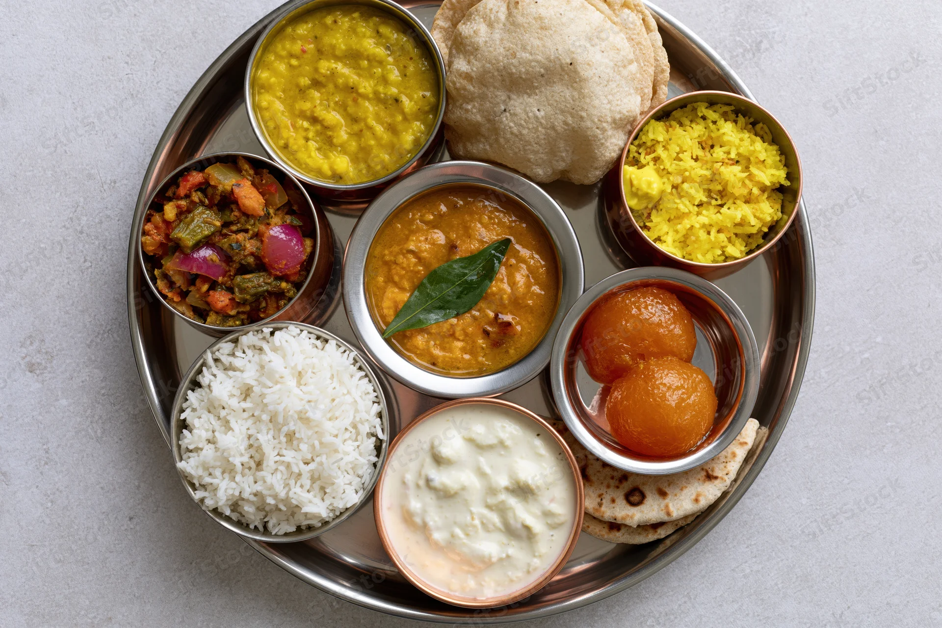 Indian thali meal served on a large steel plate with multiple small bowls of curry, rice, dal, yogurt, roti, papad, and sweet dessert arranged in a circular pattern on a light gray surface