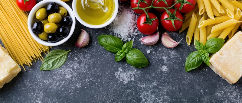 Italian cooking ingredients arranged along the bottom edge of a dark countertop with copyspace in center