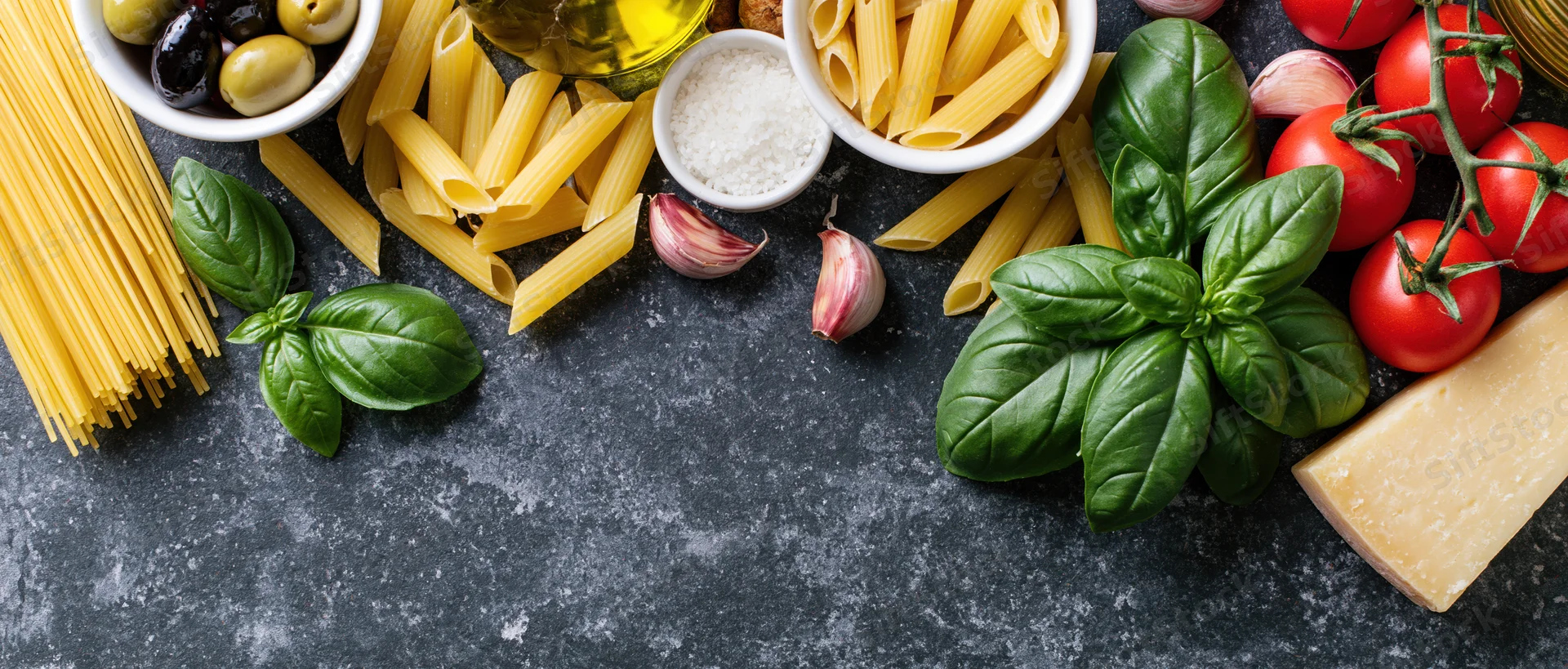 Italian cooking ingredients arranged along the bottom edge of a dark stone countertop with copyspace in the center
