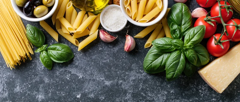 Italian cooking ingredients arranged along the bottom edge of a dark stone countertop with copyspace in the center